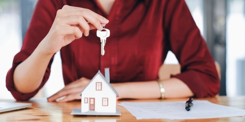 Woman in red shirt at a table holding a key above a small mini model house considering buying a house and pros and cons..